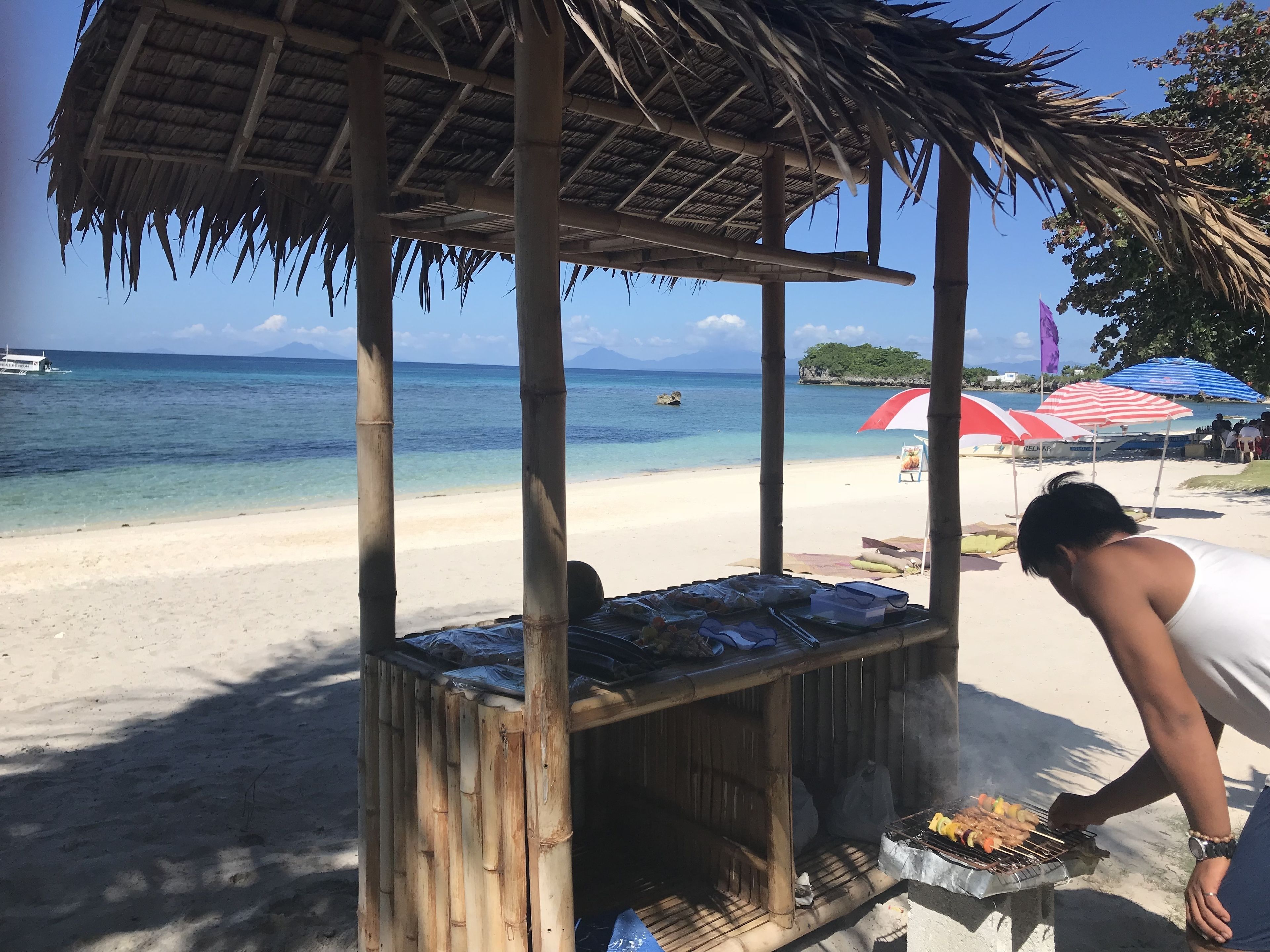 A beachside barbecue setup with a bamboo structure, a grill, and a person cooking.