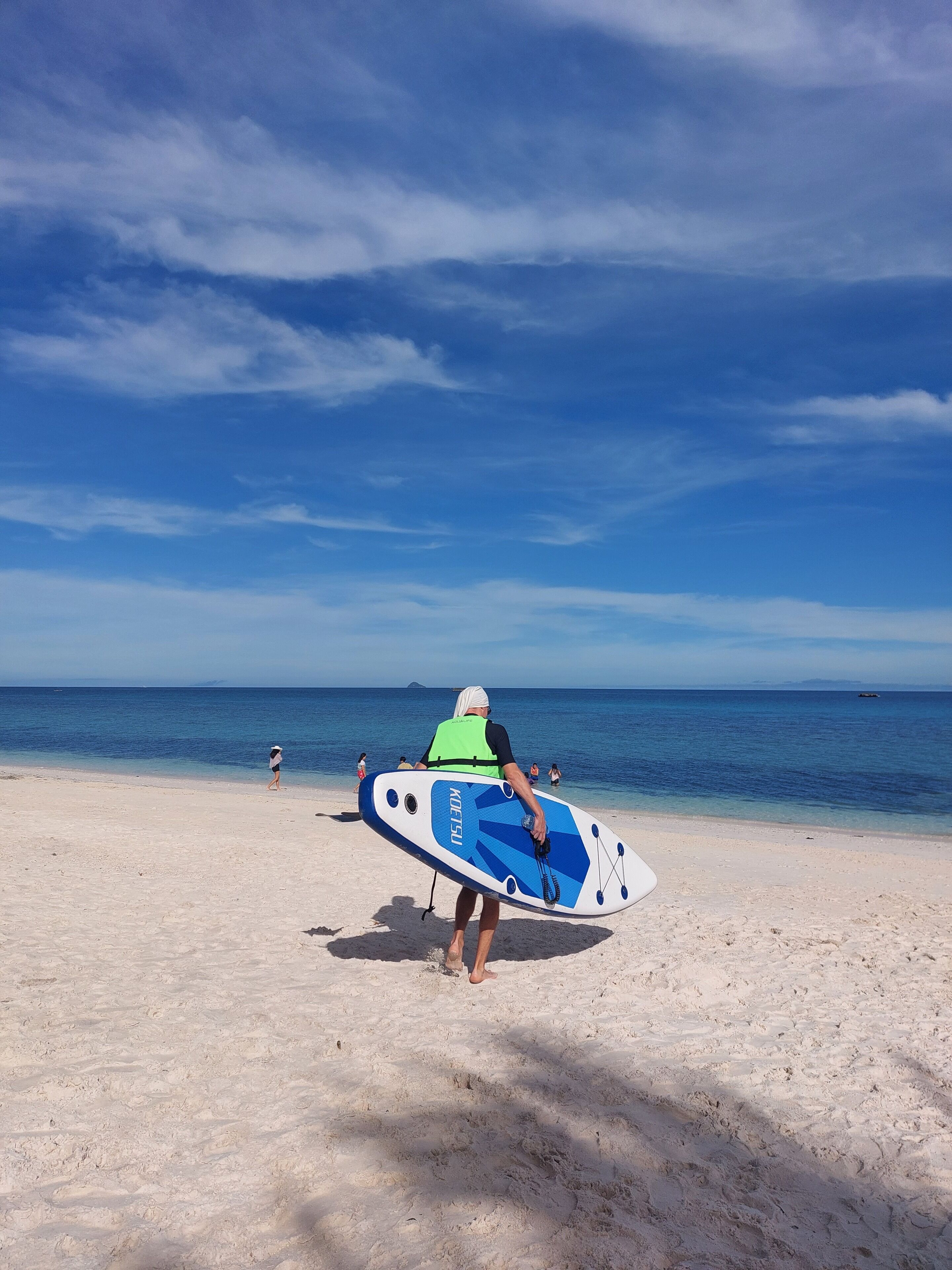 Une personne portant une planche de surf bleue et blanche sur une plage de sable, sous un ciel dégagé.