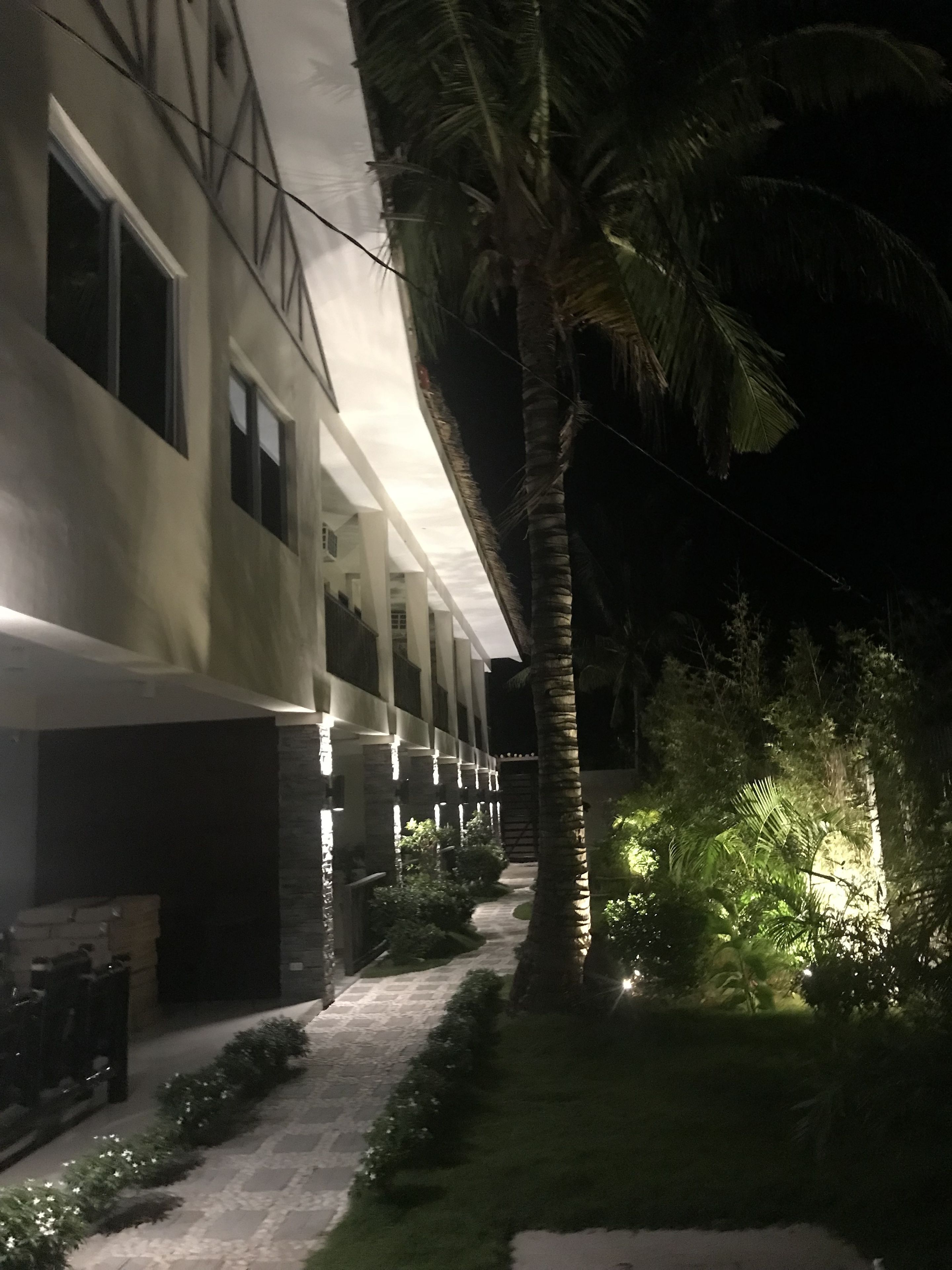 A building with a pathway leading to a gated entrance, surrounded by palm trees and greenery.