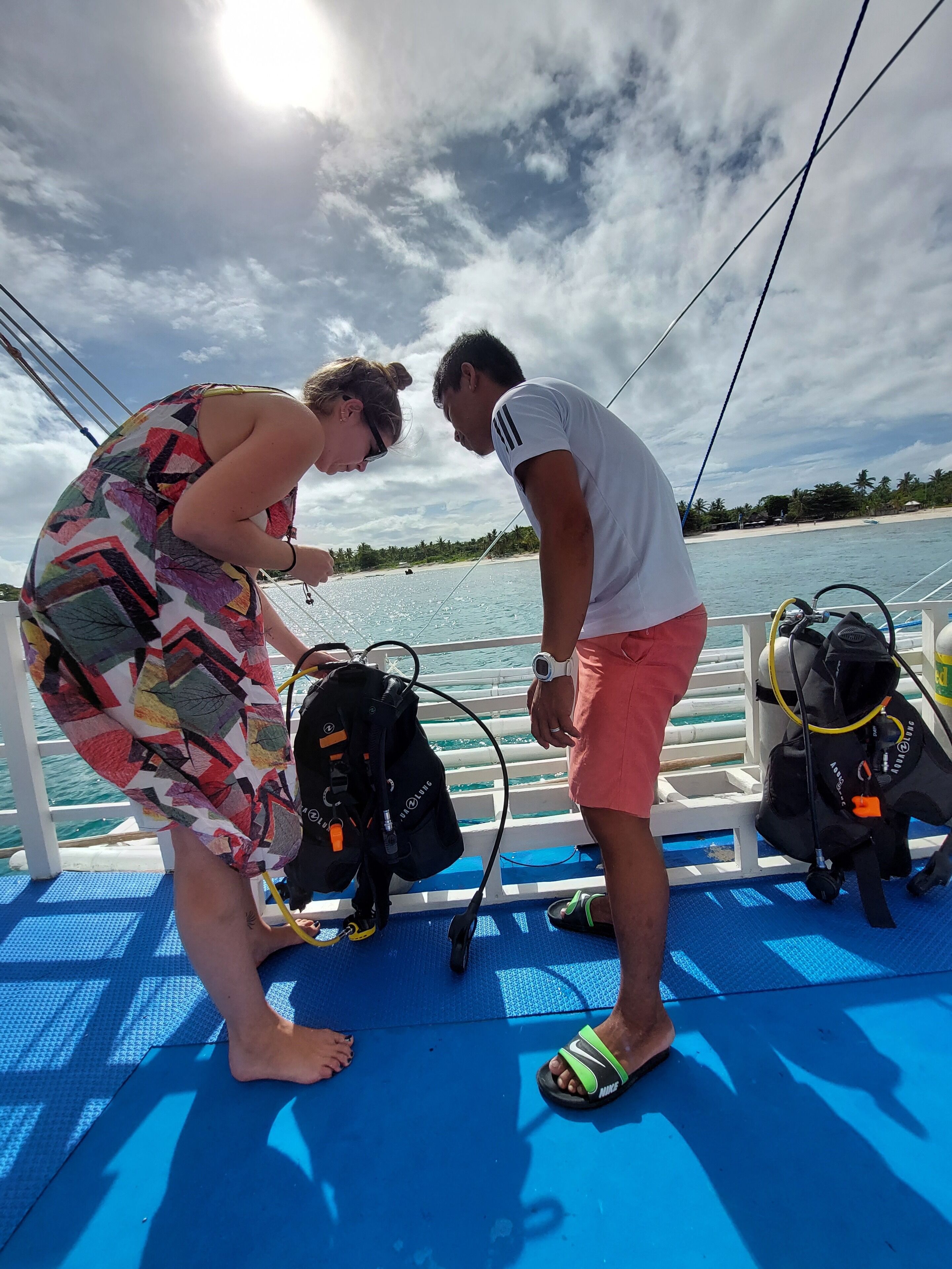 Zwei Personen bereiten Tauchausrüstung auf einem Bootsdeck vor, mit Blick auf einen Strand und Wasser.