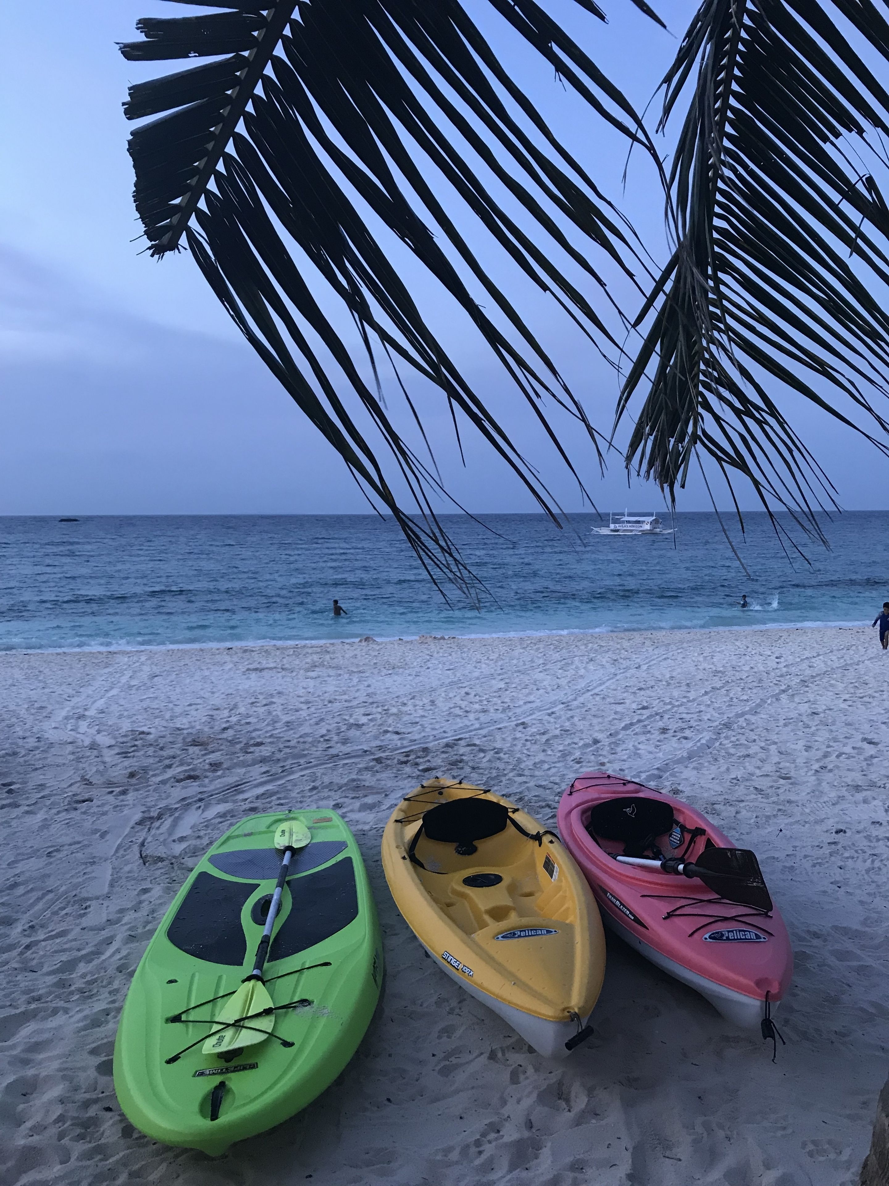 Tres kayaks sobre una playa arenosa, con vista al océano y un barco a lo lejos.