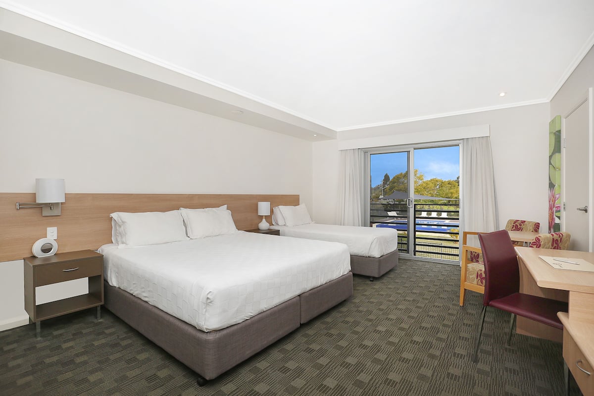 A hotel room with two beds, a desk, a chair, a nightstand, and a view of the outdoors.