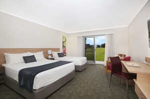 A hotel room with two beds, a desk, and a chair. There is a large window with a view of a green field.