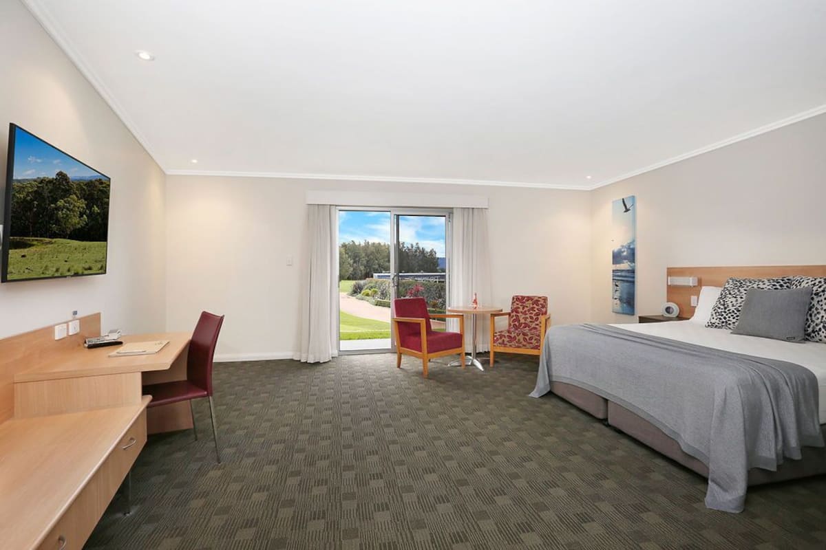 A hotel room with a bed, desk, and chairs, a large window with a view of greenery, and a painting on the wall.