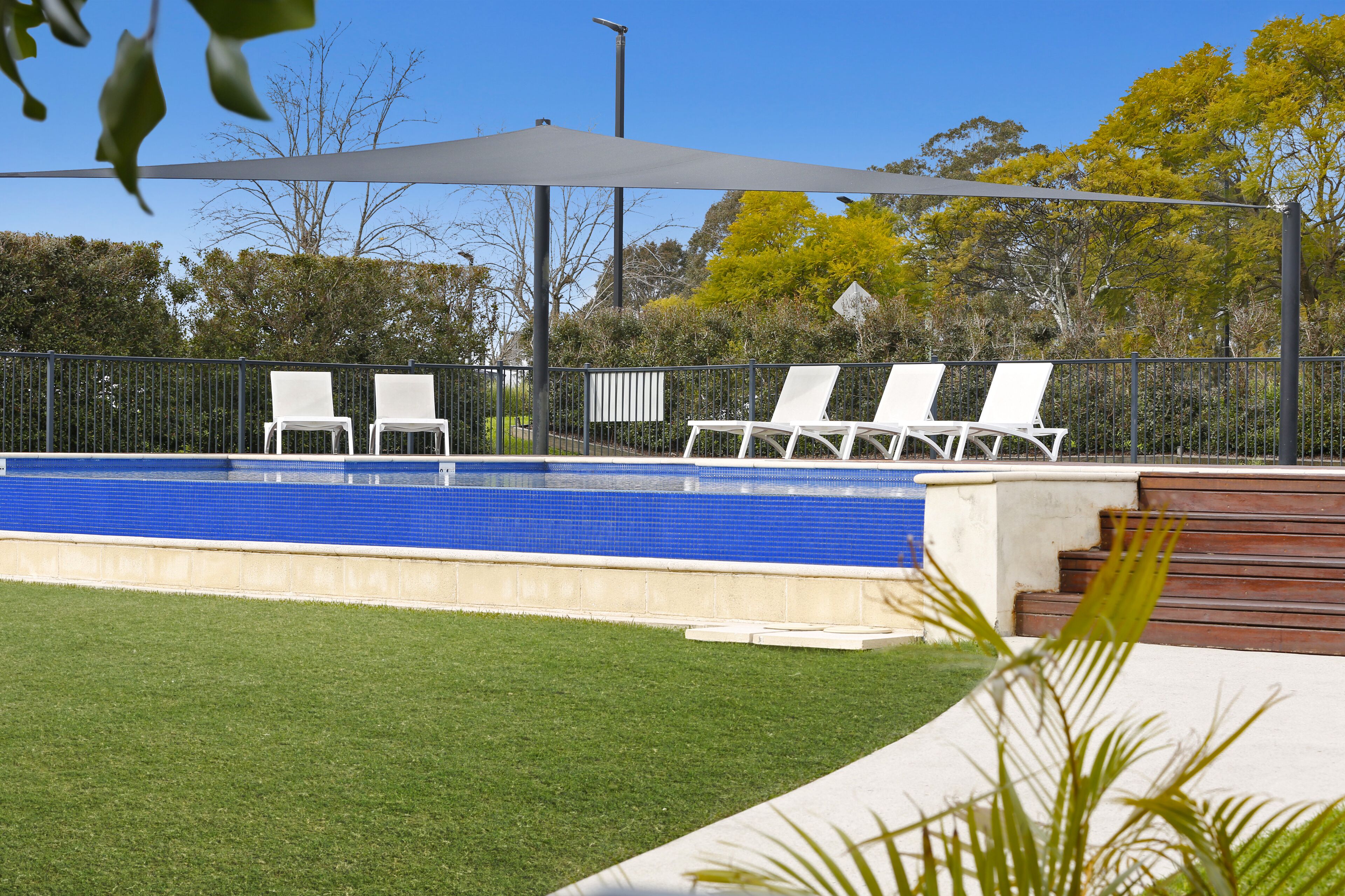 A swimming pool with three white lounge chairs under a shaded area, surrounded by a fence and greenery.