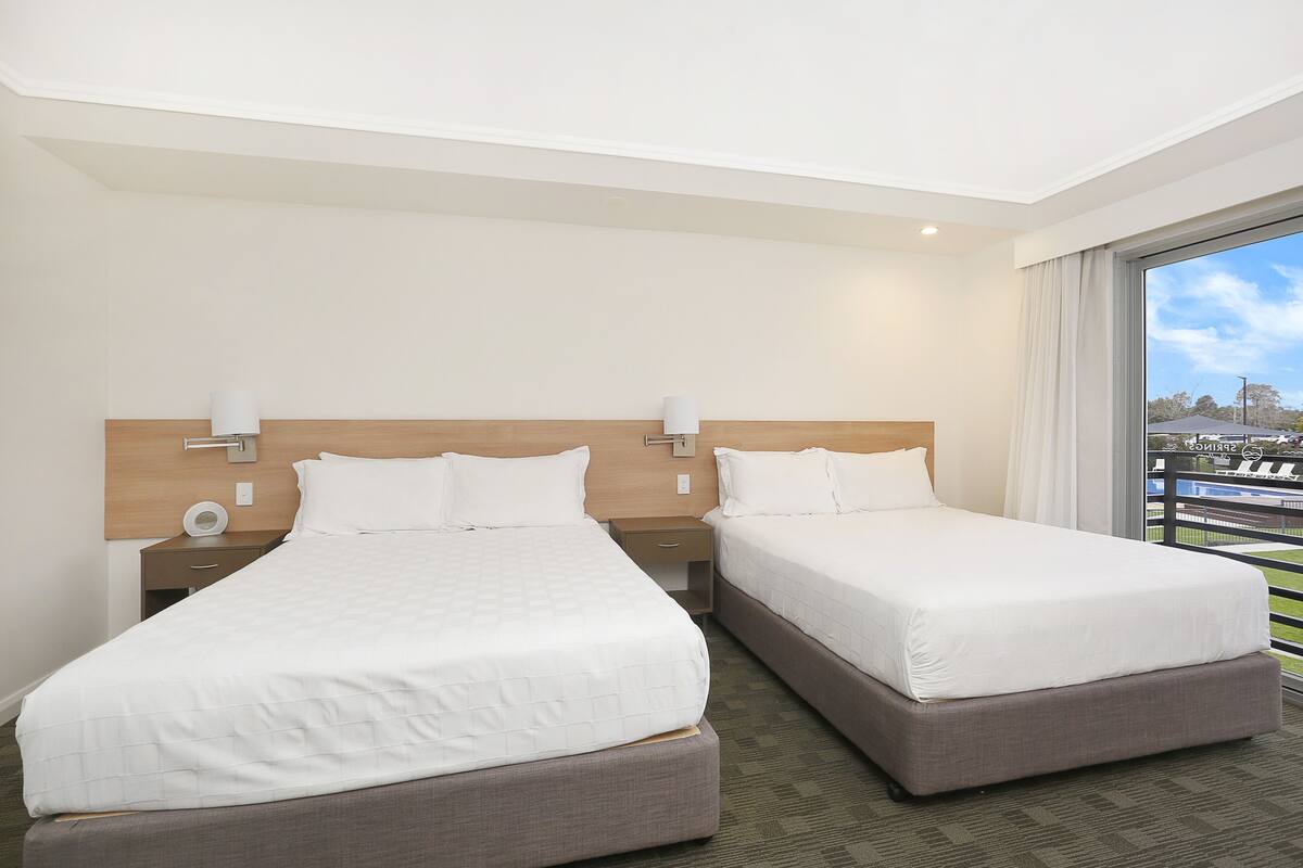 A hotel room with two beds, a wooden headboard, a nightstand with a lamp, and a large window with a view of the city.