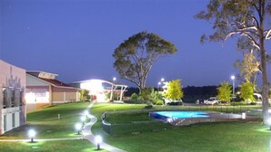 A well-lit outdoor area with a swimming pool, a building with a red roof, and a large tree. - Springs  Shoalhaven (Worrigee)
