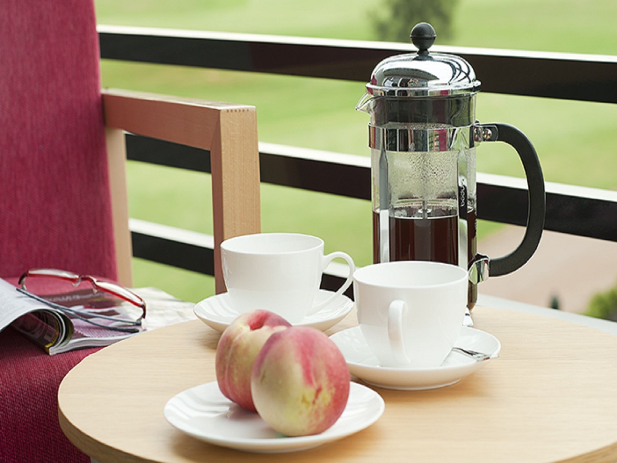 Una mesa con una cafetera, tazas y dos melocotones en un balcón con vistas a un campo verde.