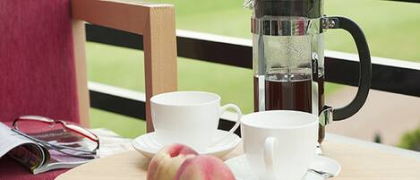 A table with a coffee pot, cups, and two peaches on a balcony with a view of a green field.