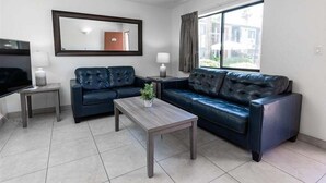A modern living room with a dark leather sofa, a wooden coffee table, a flat-screen TV, and a large window with curtains.