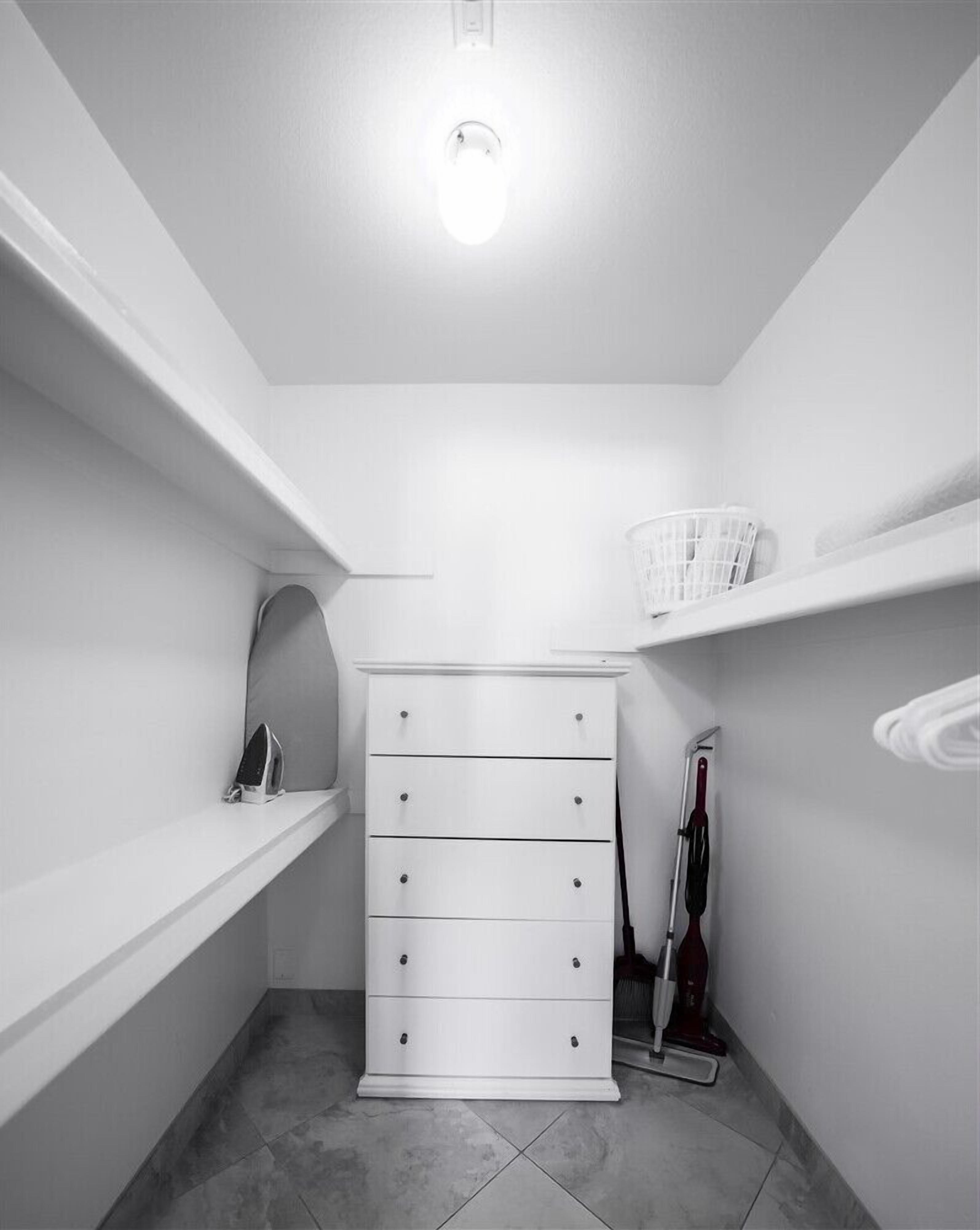 A small, white, tiled closet with a white dresser, shelves, and a broom.