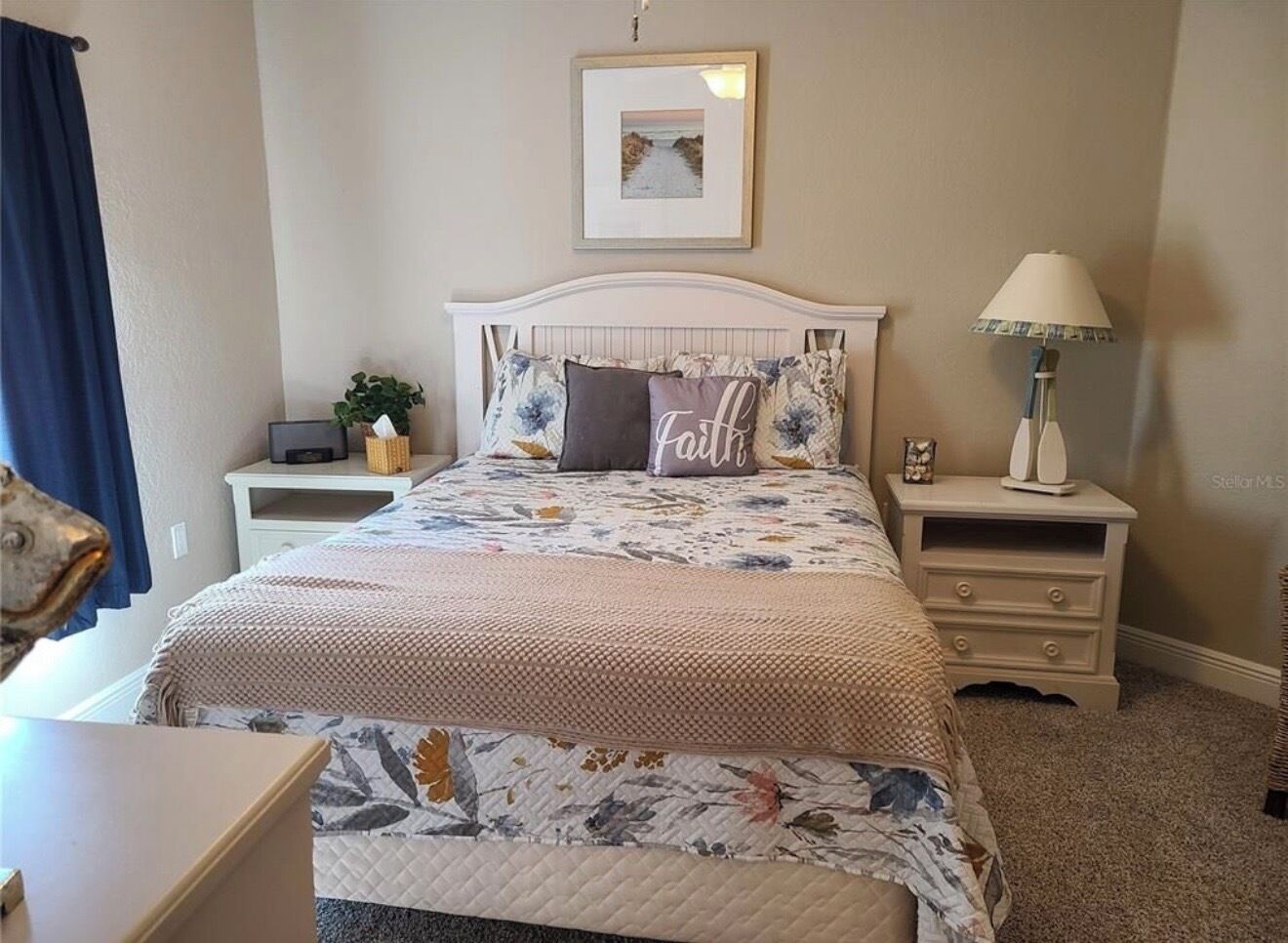 Une chambre à coucher avec un lit, deux tables de chevet, une lampe, un tableau encadré au mur et un rideau.