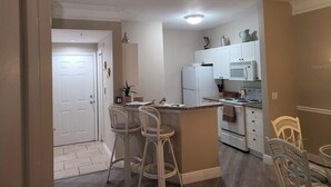 Een compacte keuken met witte apparaten, een klein kookeiland met barkrukken en een deur naar een andere kamer.