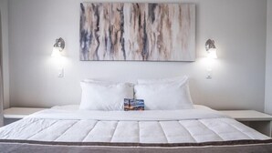 Un lit bien fait, avec une couette blanche, deux lampes de chevet et une grande peinture abstraite accrochée au-dessus.