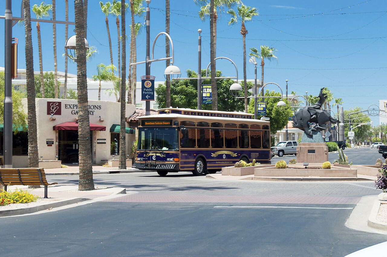 Un trolebús en una intersección urbana, con palmeras y una estatua.