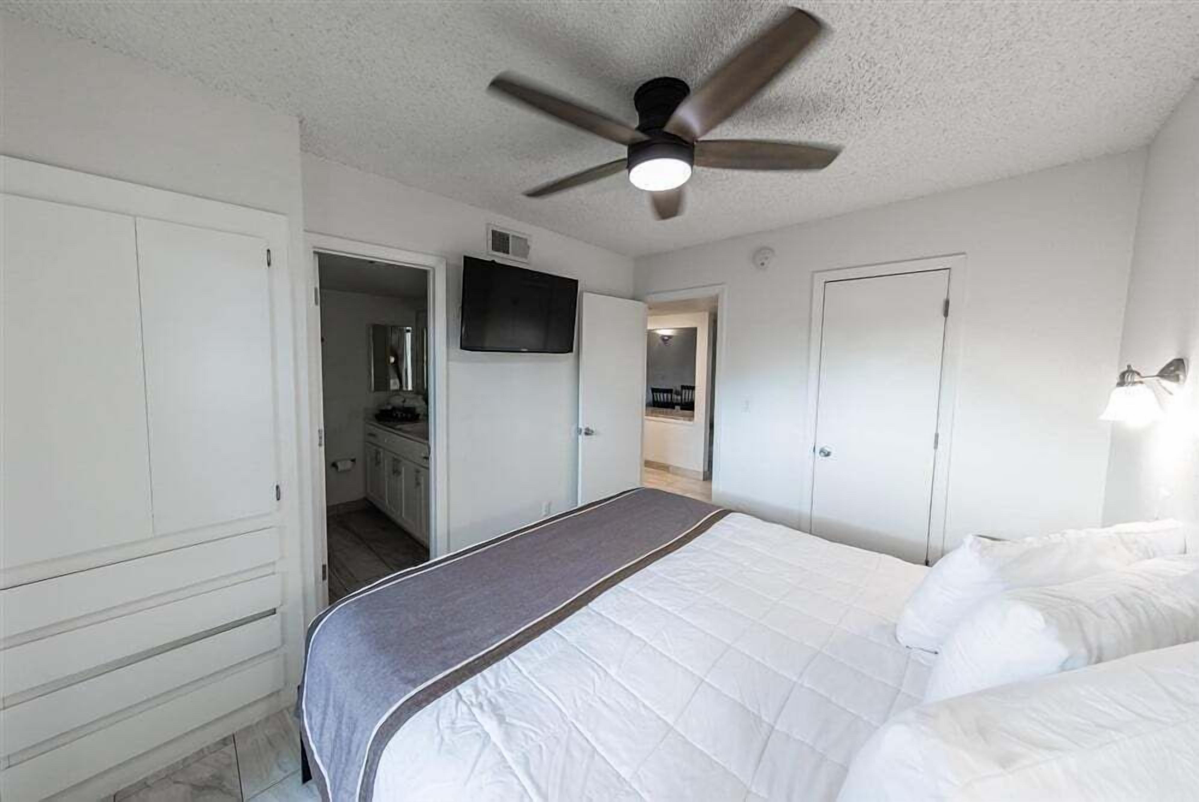 A bedroom with a bed, a ceiling fan, a TV, and a door leading to a bathroom.