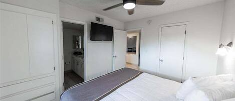 A bedroom with a bed, a ceiling fan, a TV, and a door leading to a bathroom.