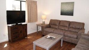 Un salon avec un téléviseur à écran plat posé sur un meuble en bois, un canapé marron, une petite table avec une lampe et un tableau accroché au mur.