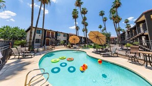 Une piscine entourée de palmiers et de bâtiments d’hôtel, avec des chaises longues et des parasols.