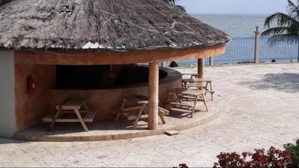 A thatched-roof outdoor seating area with a grill, surrounded by palm trees and a view of the ocean. - Amara Suites Banana Island (Lagos)