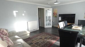 A modern living room with a glass dining table, a sofa with plaid cushions, a patterned rug, and a TV on the wall. - Huku Kwetu - Brentwood Townhouse (Dunstable)