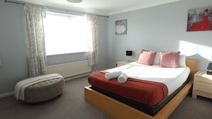 A bedroom with a large bed, a nightstand, a lamp, a chair, and a window with curtains.