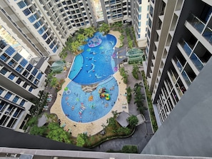 A high-rise building with multiple balconies, a central swimming pool area with various water play structures, and surrounding greenery.