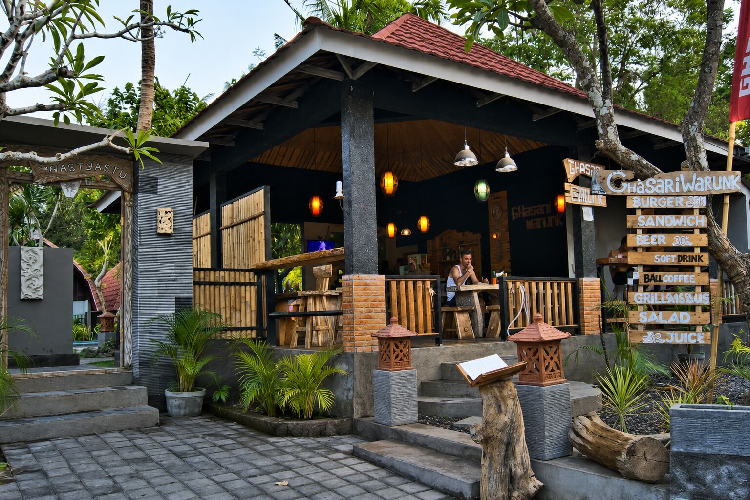 A restaurant with a covered outdoor seating area, a signboard listing food and drink options, and a person sitting at a table.
