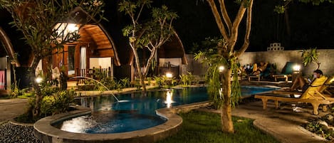 Uma área de piscina à noite, com espreguiçadeiras e um pequeno edifício com telhado curvo.