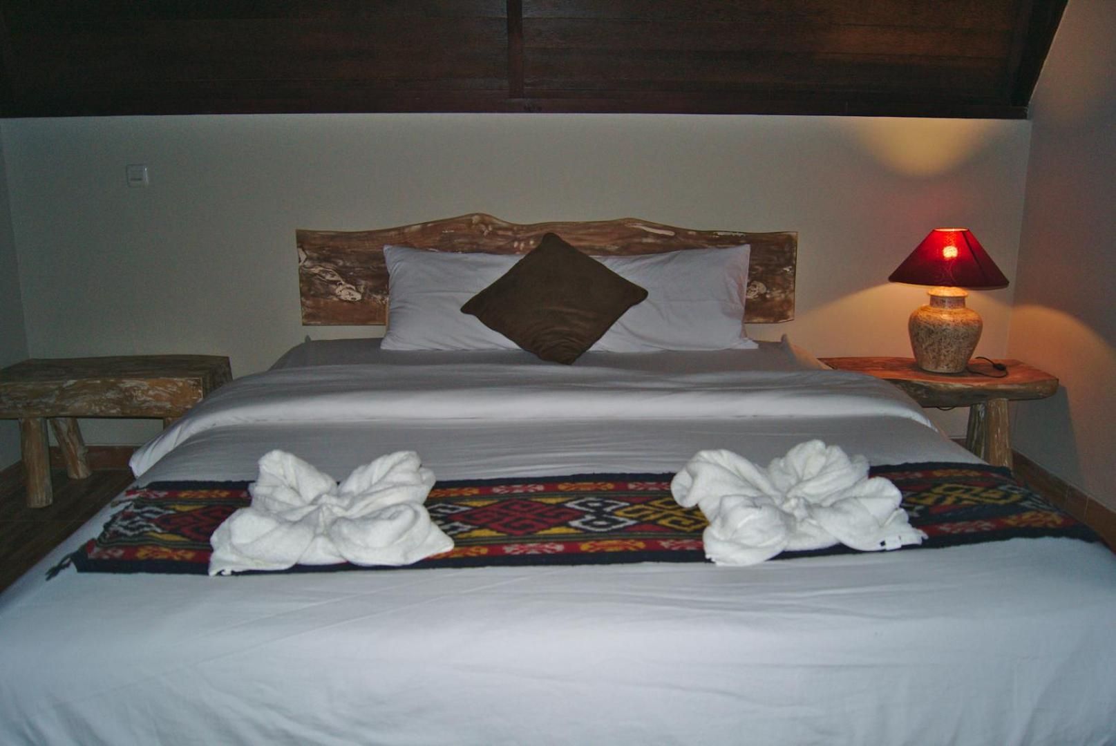 A neatly made bed with white linens, a brown headboard, and a red lamp on a wooden nightstand.