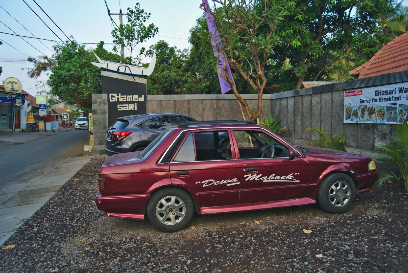 A maroon car parked on a gravel surface with a sign in the background.