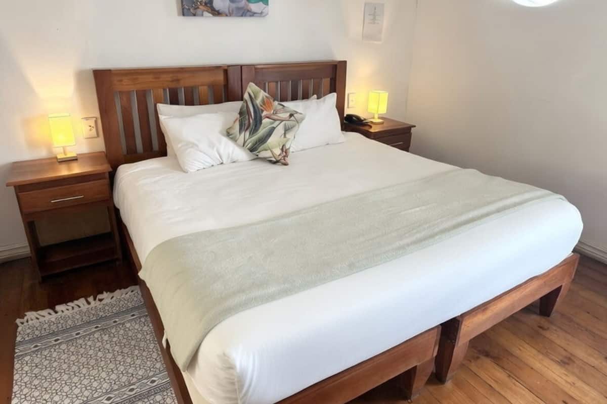 Ein ordentlich bezogenes Bett mit Holz-Kopfteil, zwei Nachttische mit Lampen, ein gemustertes Teppich und eine Wand mit einem gerahmten abstrakten Gemälde.