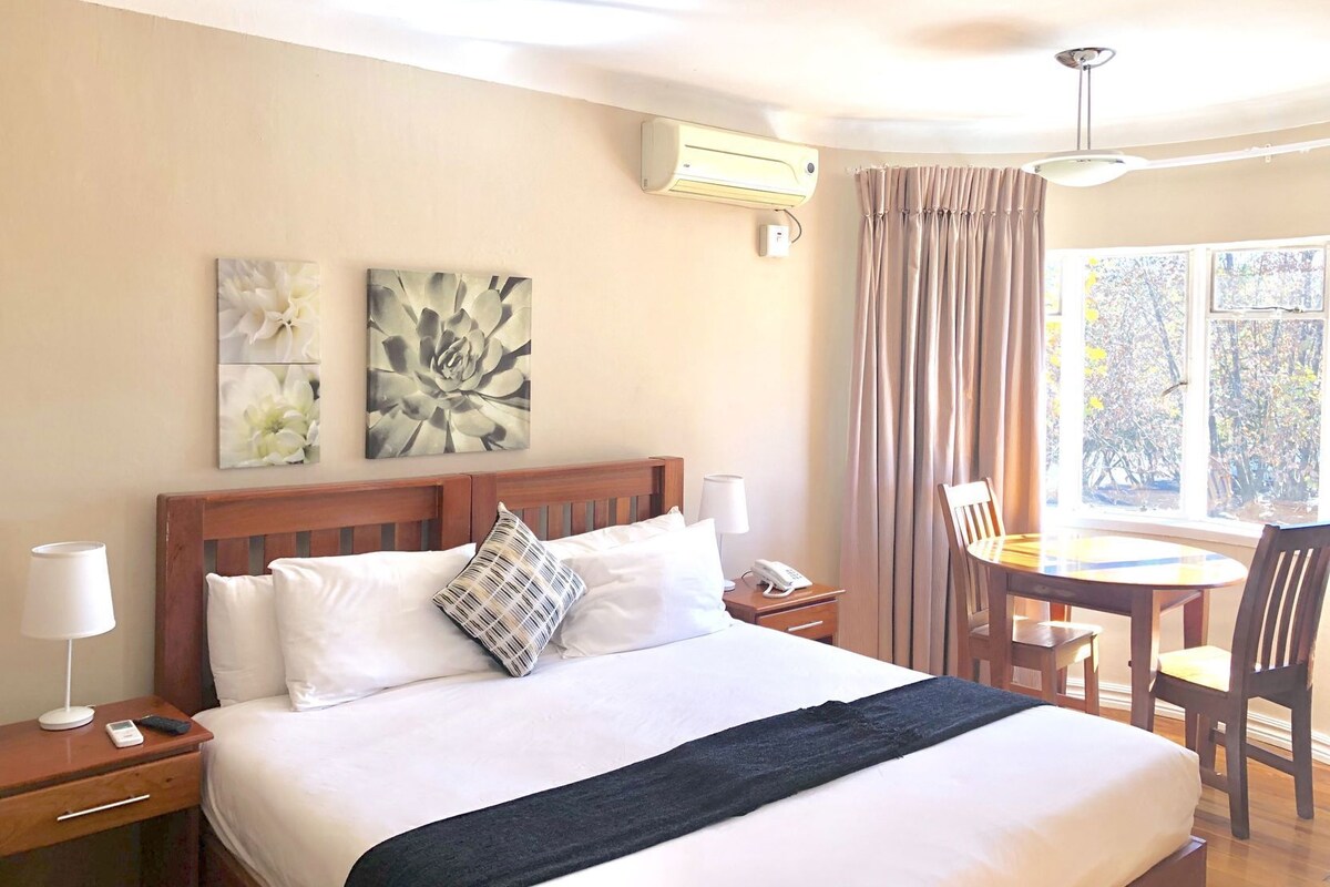 Une chambre d’hôtel comprenant un lit, des tables de chevet, un bureau avec des chaises, une fenêtre avec des rideaux et un climatiseur fixé au mur.