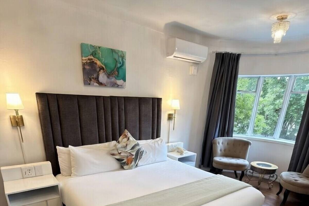 Ein großes Bett mit dunklem Kopfteil, weißer Bettwäsche und einem gemusterten Teppich. Ein Nachttisch mit Lampe, ein Sessel und ein Fenster mit Vorhängen.