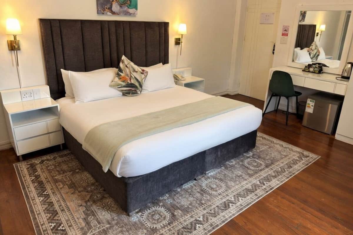 Ein ordentlich eingerichtetes Hotelzimmer mit einem großen Bett, zwei Nachttischen, einem Schreibtisch und einem Spiegel.