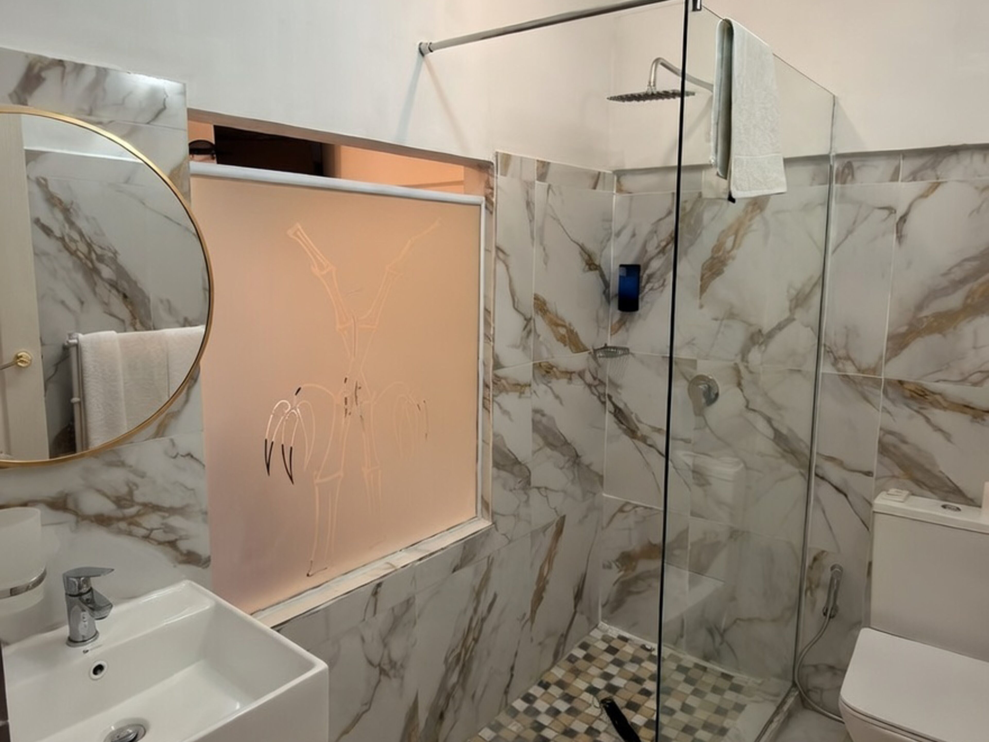 Un baño con pared de mármol, cabina de ducha de vidrio, lavamanos blanco y retrete.