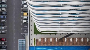 Vue aérienne d’un bâtiment moderne à la façade ondulée originale, d’une piscine sur laquelle on peut lire les mots « THE WAVE », et d’un parking avec des voitures.