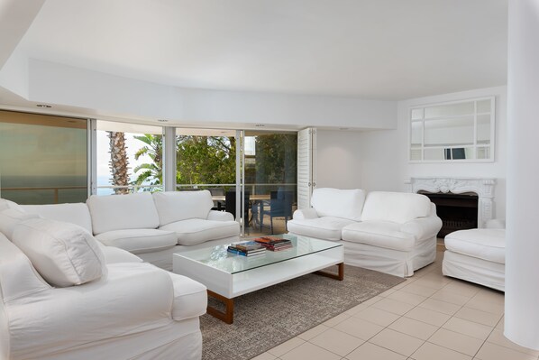 A modern living room with white furniture, a glass coffee table, and a fireplace.