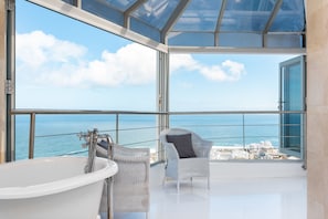 A modern bathroom with a large bathtub, a wicker chair, and a balcony with a view of the ocean and cityscape.