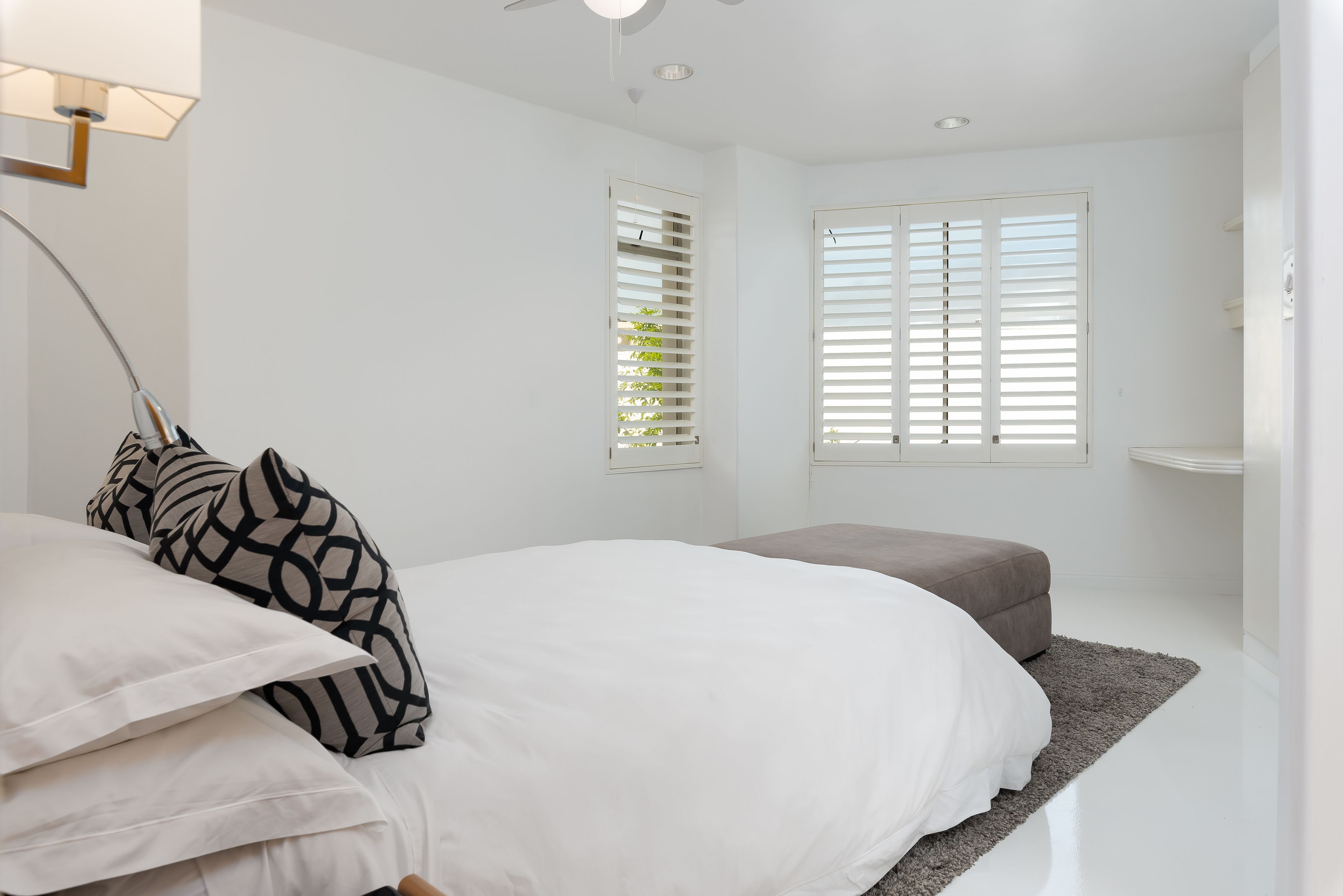 A bedroom with a large bed, a bedside table, a grey bench, and two windows with blinds.