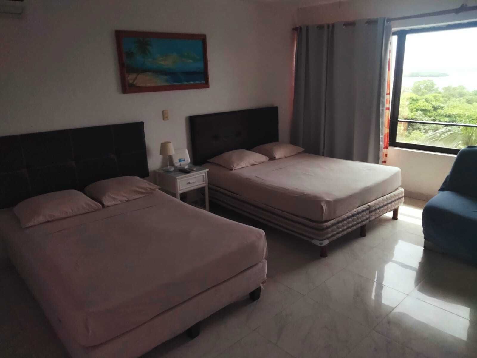 A hotel room with two beds, a nightstand, a painting on the wall, and a view of greenery through the window.