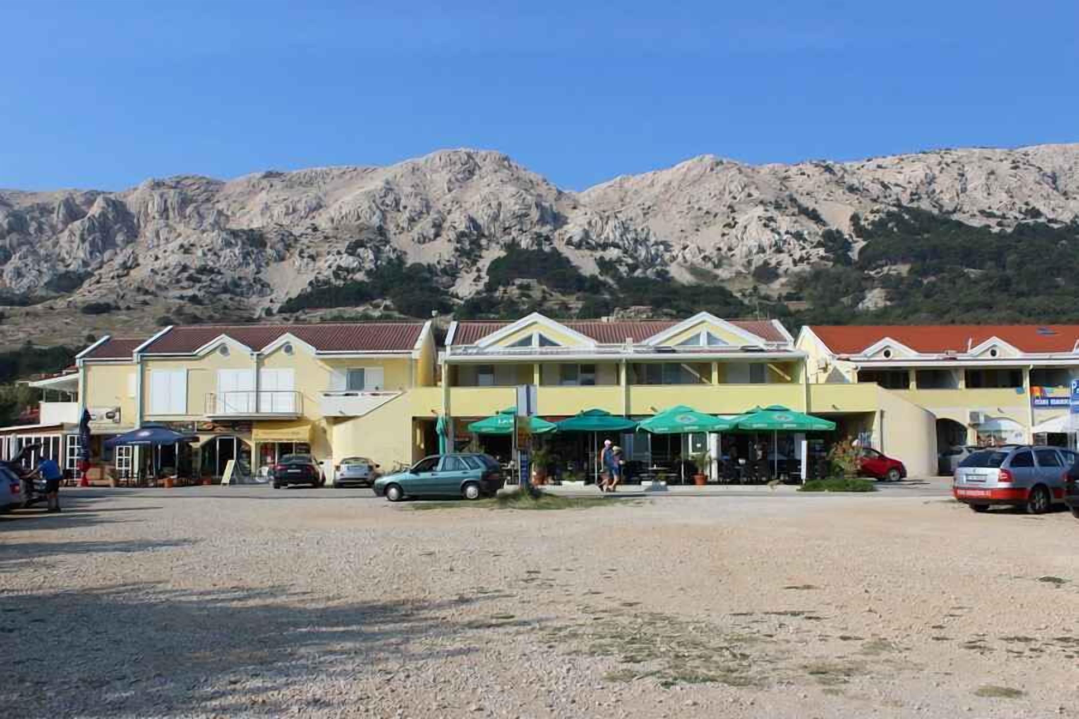 A row of yellow buildings with red roofs, a parking area, and a mountainous backdrop.