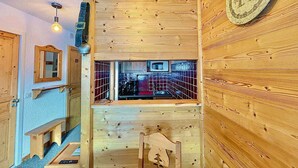 Un intérieur en bois avec un coin cuisine visible à travers une fenêtre, une horloge au mur et un banc près de l’entrée.