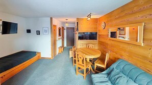 Un espace de vie compact comprenant un canapé, une table à manger avec des chaises, un coin cuisine et un téléviseur fixé au mur.