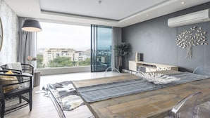 Ein modernes Wohnzimmer mit einem Holzesstisch, transparenten Glasstühlen, einem schwarzen Sofa und einem großen Fenster mit Blick auf die Stadt.