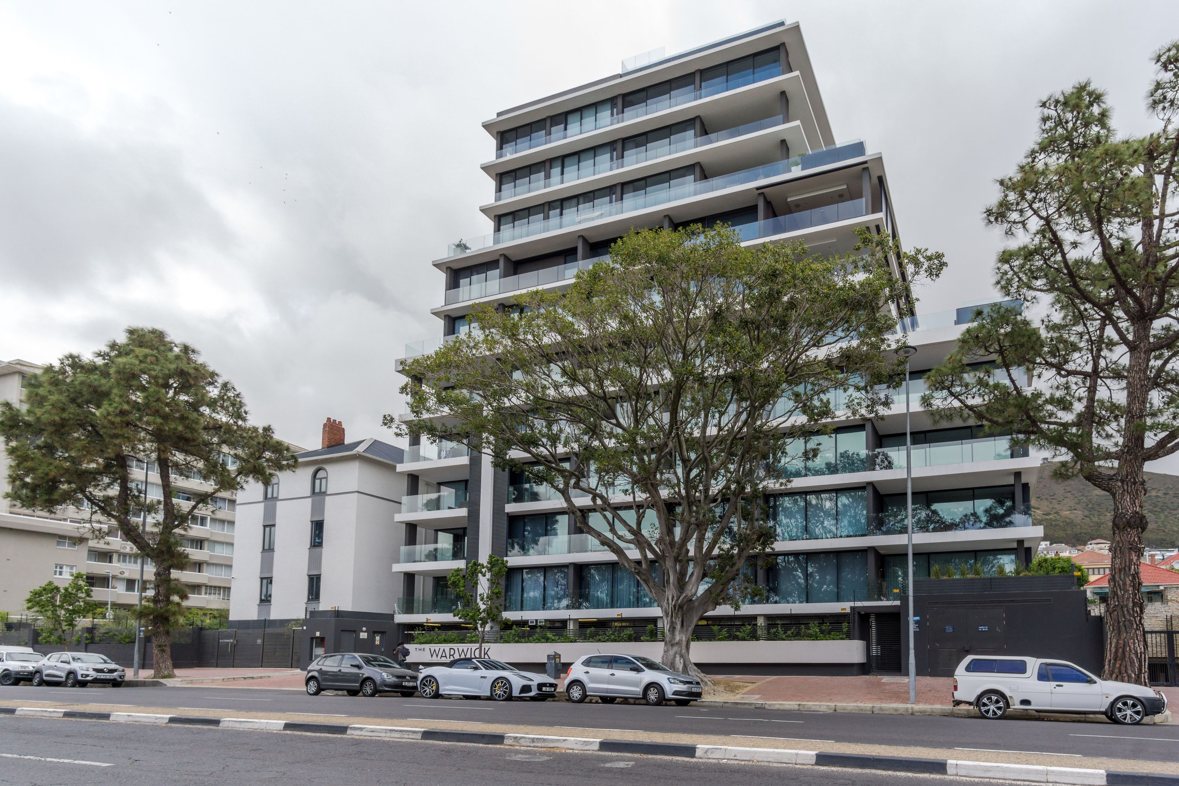 A modern multi-story building with a flat roof, large windows, and a parking area with several cars.