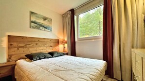 A bedroom with a wooden headboard, a bed with white bedding, a nightstand with a lamp, a dresser, and a window with curtains.