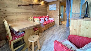 A cozy cabin interior with a wooden wall, a wooden table, a sofa with red cushions, and a small stool.
