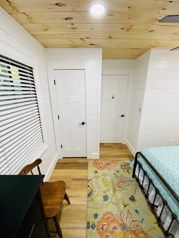 Una habitación con una cama, un escritorio y una silla. Hay dos puertas y una ventana con persianas.