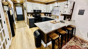 Una cucina moderna con mobili bianchi, un piano cottura nero e un ripiano in marmo. Sono presenti sgabelli in legno e un cestino.